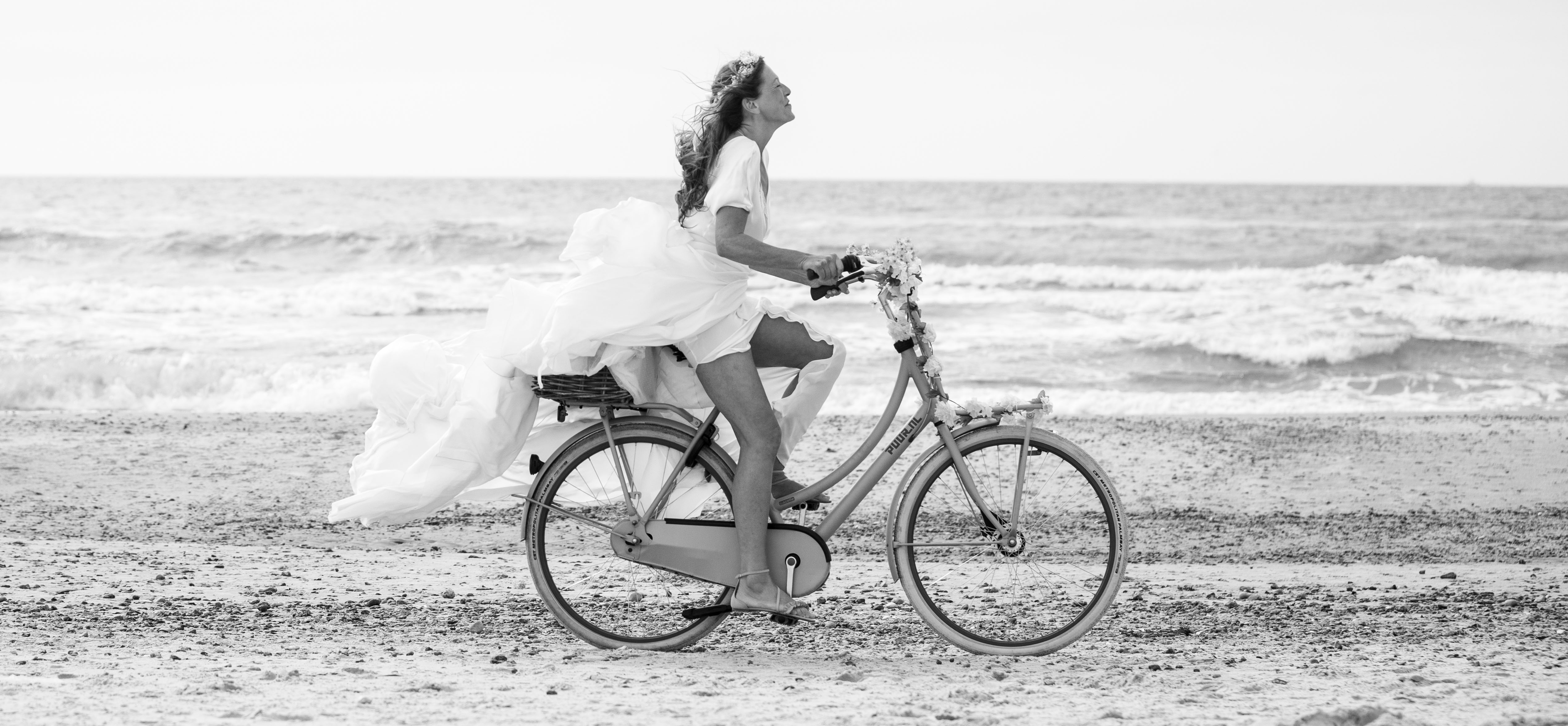 Glad kvinde i bryllupskjole cykler på stranden på en cykel