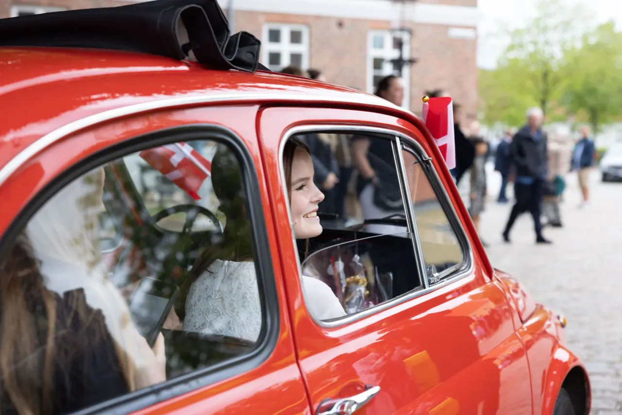 Smilende konfirmant sidder i sød rød lille bil
